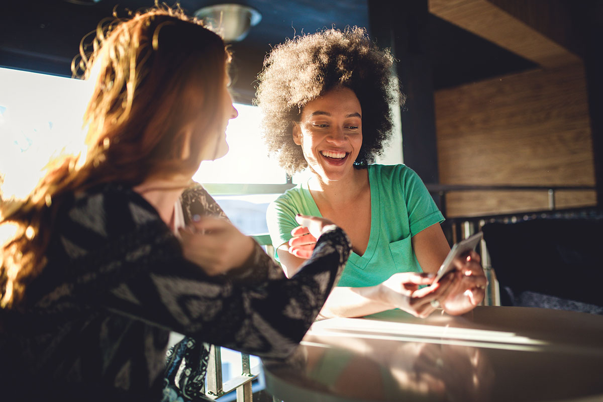 Two women sit in a cafe looking at their smart phones.