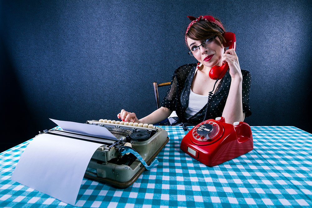 Woman talking on a rotary telephone sitting at table with typewriter